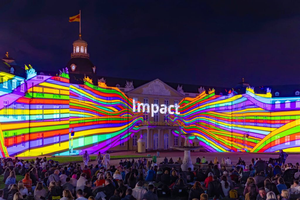 Lichtinstallation auf Schlossfassade bei Nacht, zeigt bunte Streifen und das Wort "Impact", Publikum sitzt davor. Passend für lokale Werbung.